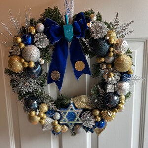 May include: A blue and silver Christmas wreath with a blue velvet bow and a blue dreidel hanging in the centre. The wreath is decorated with gold and silver baubles, pine cones, and sprigs of greenery.