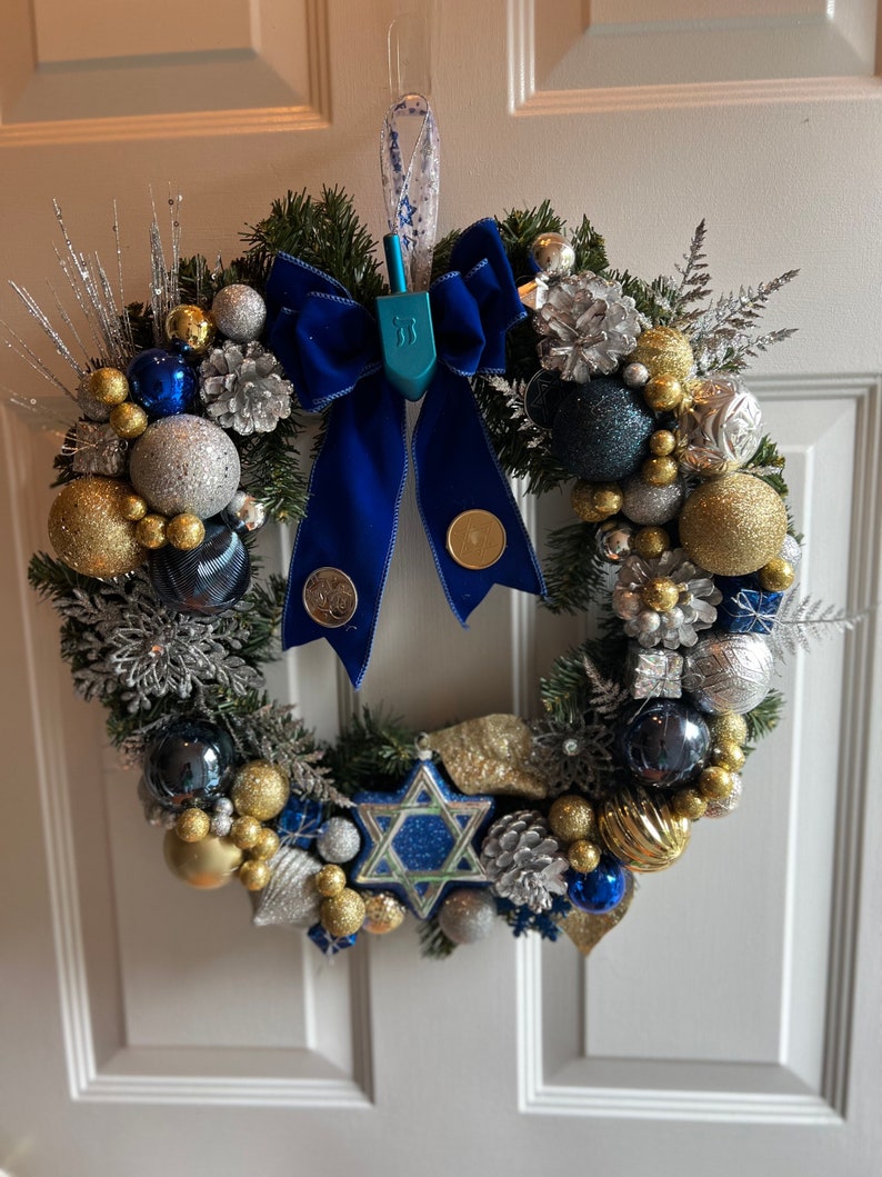 May include: A blue and silver Christmas wreath with a blue velvet bow and a blue dreidel hanging in the centre. The wreath is decorated with gold and silver baubles, pine cones, and sprigs of greenery.