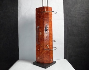 Könnte beinhalten: Eine hohe, zylindrische Skulptur aus Holz mit einer braunen, strukturierten Oberfläche. Die Skulptur hat einen Metallgriff und einen Metallring oben. Die Skulptur steht auf einem schwarzen Sockel.