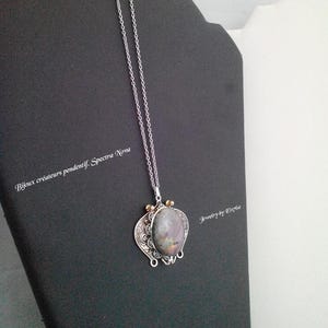 May include: A silver necklace with a large, oval pendant. The pendant features a grey and purple stone set in an ornate, filigree silver frame with gold accents. The necklace is displayed on a black jewellery stand. Text on the image reads "Bijoux créateurs pendentif. Spectra Nova" and "Jewelry by Elpize".
