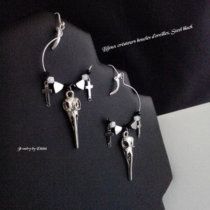 May include: Steel black earrings with a gothic theme. The earrings feature a bird skull pendant, small crosses, and black and white beads. The text "Bijoux créateurs boucles d'oreilles. Steel black" is visible.