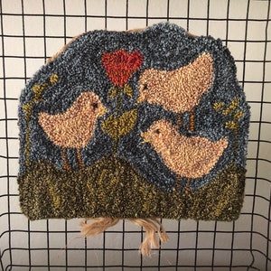May include: A hand-stitched rug featuring three yellow chicks with red beaks and legs, standing in front of a red flower on a blue background. The chicks are surrounded by green foliage.
