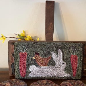 May include: A white rabbit with a brown chicken on its back, two red carrots, and green foliage are depicted in a needlepoint design on a wooden frame.