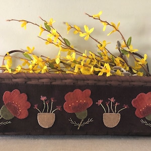May include: A brown wicker basket with a felt panel featuring three red embroidered flowers in pots. The basket is filled with yellow forsythia branches.
