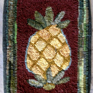 May include: A close-up of a hand-stitched rug with a pineapple design. The pineapple is yellow and green, and the rug is surrounded by a brown border with green and blue accents.