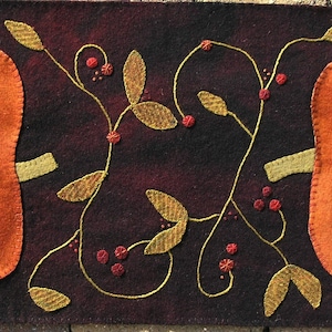 May include: A brown felt table runner with two orange felt pumpkins and a vine with green leaves and red berries.