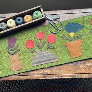 May include: A green wool runner with appliqued flowers in pots. A wooden box holds spools of yarn in various colors. A pair of scissors rests on the runner, suggesting a craft project.