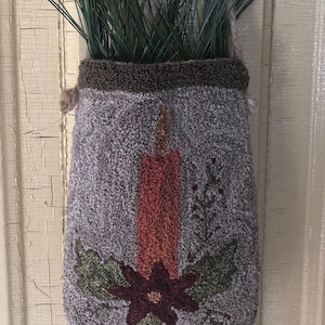 May include: A grey and brown wool wall hanging with a red candle and a burgundy flower with green leaves. The hanging is decorated with a brown rope and has a green plant hanging from the top.