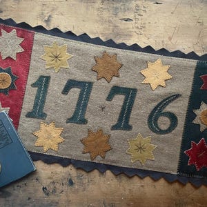 May include: A rectangular felt wall hanging featuring the year 1776 in dark green stitched numbers. The center is tan with yellow and brown star appliques. The left border is red with stars, and the right border is dark green with red and yellow stars.