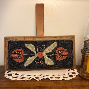 May include: A vintage wooden rug beater with a floral design. The rug beater features a dark background with orange and green floral elements. A small oil lamp with a glass chimney sits to the right of the rug beater.