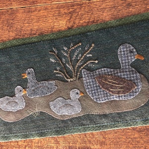 May include: A green felt wall hanging with a family of ducks. The mother duck is larger than the three ducklings. The ducks are made of felt and have a plaid pattern. The background is a dark green felt with a small brown felt bush.