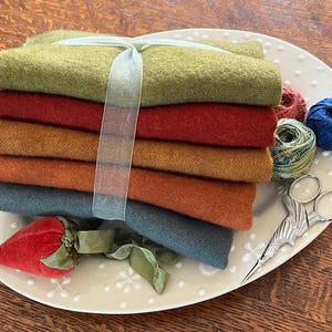 May include: A stack of colorful wool fabric squares tied with a sheer ribbon, displayed on a white oval plate. The stack includes shades of green, red, orange, and blue. Also on the plate are spools of thread and decorative scissors.