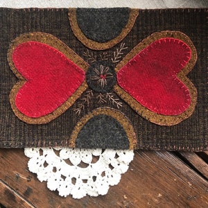 May include: A brown wool felt wall hanging with a red felt heart design. The heart is surrounded by a brown felt border and a brown felt background. The heart is stitched with a brown thread.