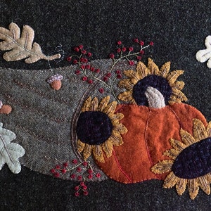 May include: A felt appliqué wall hanging with a fall theme. It features a pumpkin with sunflowers, oak leaves, and acorns in shades of brown, orange, yellow, and green.