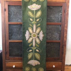 May include: A green fabric wall hanging with a floral design. The design features three stylized flowers with white and yellow petals and green leaves. The background is a dark green color.