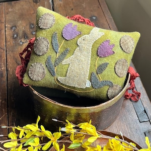 Könnte beinhalten: Ein grünes Filzkissen mit einem weißen Hasen, lila Blumen und braunen Kreisen. Das Kissen liegt auf einer Holzkiste.