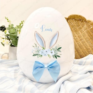 May include: A white ceramic Easter egg decorated with a blue bow, bunny ears, and floral accents. The egg is personalised with the name "Louis".