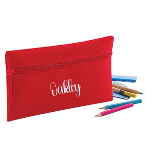 Personalised classic red school pencil case with secure zip fastening and custom name in white. Shown with pencils, ruler and school stationery to demonstrate everyday use. Durable 600D polyester pencil pouch with 0.5 litre capacity, ideal for school, nursery and back to school essentials.
