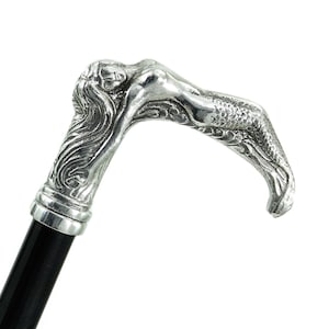 May include: A walking stick featuring a silver-toned metal handle shaped like a reclining mermaid with long flowing hair and a detailed tail. The handle is attached to a smooth, solid black shaft, creating a contrast between the metallic figure and the dark base.
