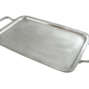Peut inclure: Un plateau de service rectangulaire argenté avec deux poignées incurvées. Le plateau a un bord légèrement surélevé et une surface polie, reflétant la lumière. Les poignées sont fixées aux petits côtés du plateau.