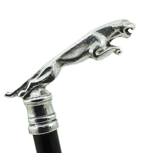 Peut inclure: Une canne de marche avec une poignée en métal argenté en forme de jaguar bondissant en position de saut. La poignée est fixée sur un bâton noir lisse, avec un collier décoratif présentant des rainures horizontales reliant les deux parties.