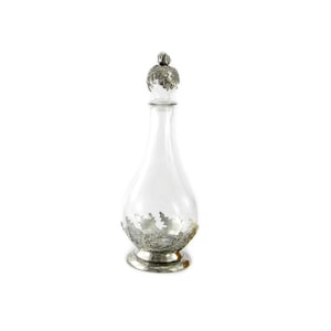 May include: A clear glass decanter with a silver-coloured metal stopper and base. The stopper and base are decorated with an intricate design of leaves.