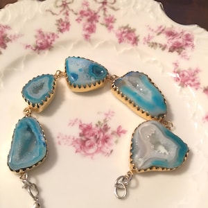May include: A bracelet featuring five irregular, blue and white geode slices with crystalline centers, each encased in a gold-toned metal setting. The segments are connected by small gold-toned jump rings, with a silver-toned lobster claw clasp.