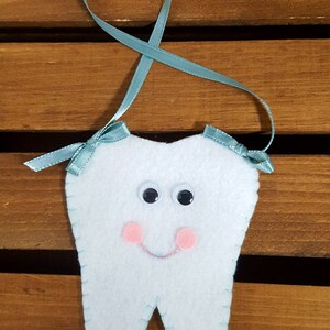 May include: A white felt tooth with googly eyes, pink cheeks, and a stitched smile. It has a light blue ribbon for hanging and small bows at the top. The tooth is a whimsical design.
