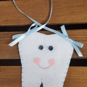 May include: A light blue felt tooth with a smiling face, pink cheeks, and black button eyes. The tooth is attached to a light blue ribbon with a bow on each end.