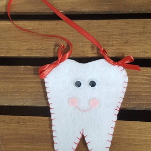 May include: A white felt tooth with a red ribbon, googly eyes, and a pink smile. The tooth is sewn with red thread.