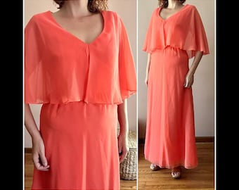 May include: A coral-coloured formal dress with a V-neckline and a flowing cape overlay. The dress has a floor-length skirt and short, flutter sleeves. The fabric appears lightweight and semi-sheer, creating an elegant silhouette.