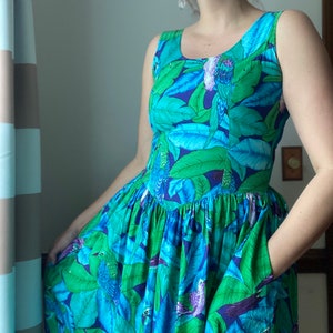 Op de afbeelding: Een maxi-jurk zonder mouwen met een blauwe en groene tropische print met vogels en bladeren. De jurk heeft een aansluitend bovenlijf en een volle rok met zakken.