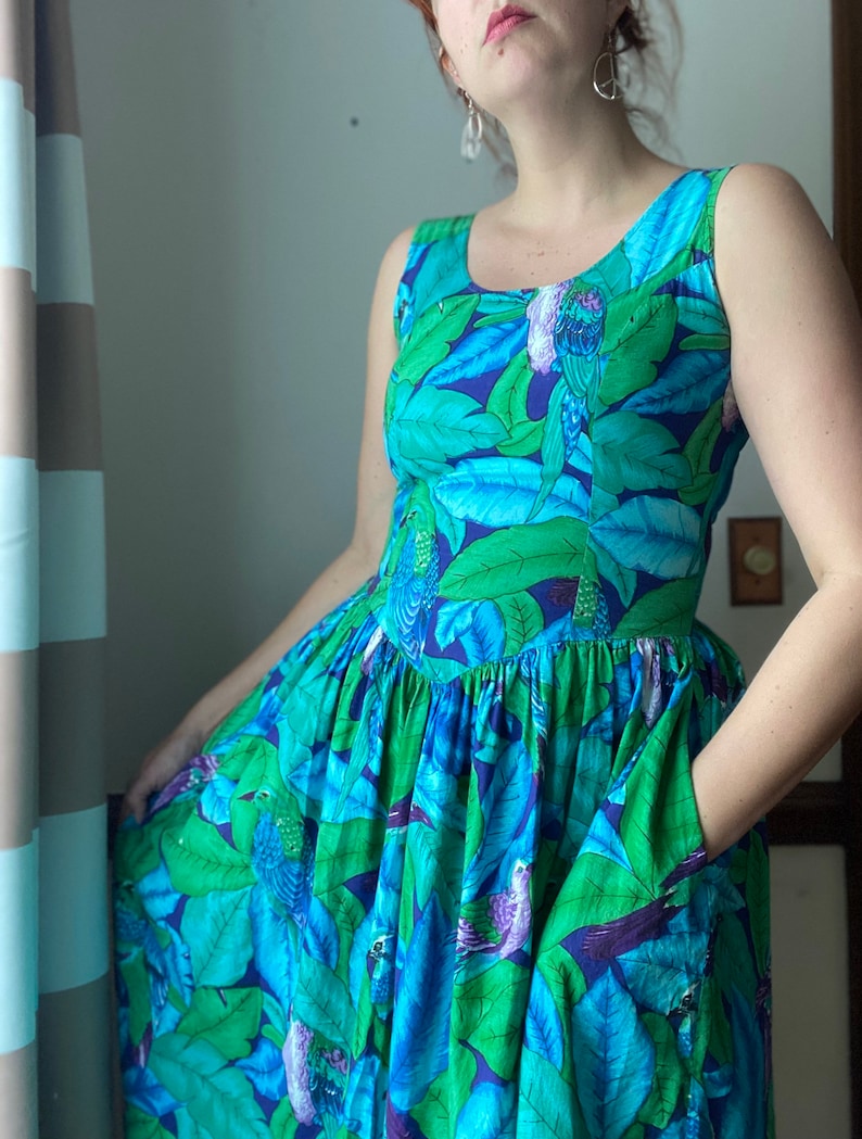 Op de afbeelding: Een maxi-jurk zonder mouwen met een blauwe en groene tropische print met vogels en bladeren. De jurk heeft een aansluitend bovenlijf en een volle rok met zakken.