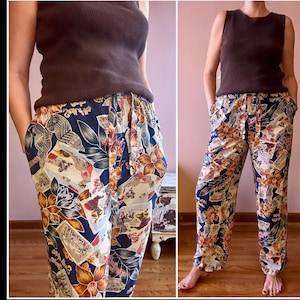 May include: Patterned trousers with a navy blue base, adorned with floral and vintage postcard motifs in orange, cream, and brown. The trousers have a drawstring waist and pockets. The person is wearing a brown vest top. The trousers are loose-fitting.