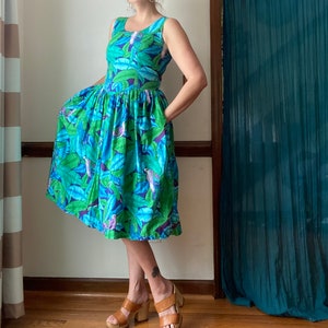 Op de afbeelding: Een blauwe en groene jurk met bloemenprint, een geplooide rok en een mouwloos lijfje. De jurk heeft een tropische print met vogels en bladeren.