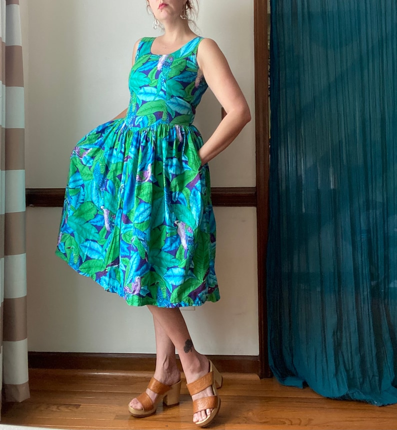 Op de afbeelding: Een blauwe en groene jurk met bloemenprint, een geplooide rok en een mouwloos lijfje. De jurk heeft een tropische print met vogels en bladeren.