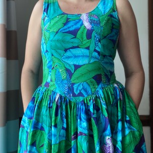 Op de afbeelding: Een mouwloze jurk met een blauwe en groene tropische print met vogels en bladeren. De jurk heeft een getailleerd bovenstuk en een volle rok.