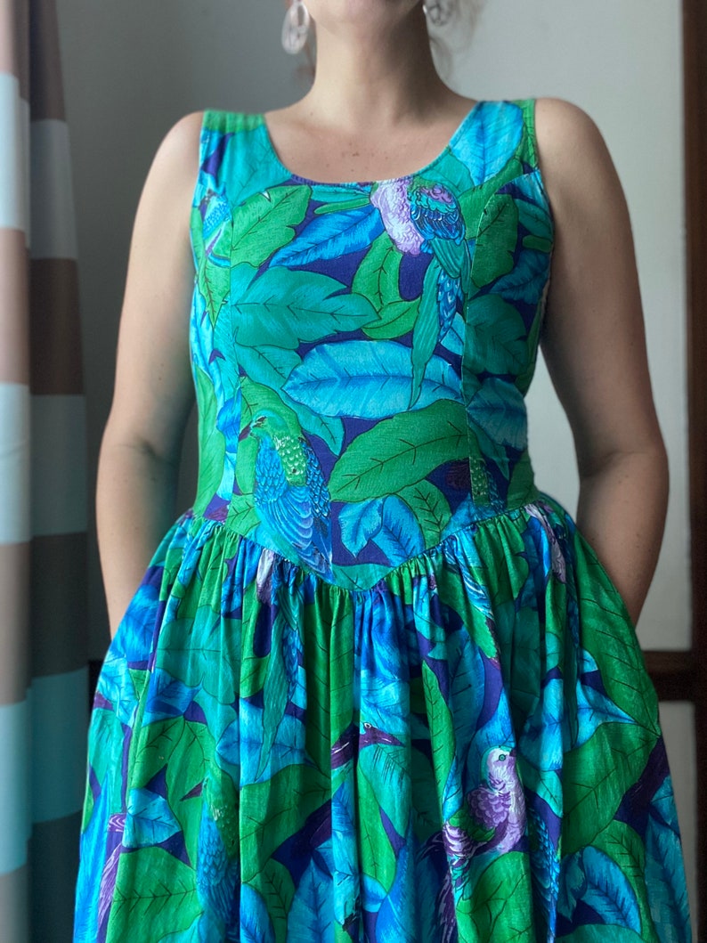 Op de afbeelding: Een mouwloze jurk met een blauwe en groene tropische print met vogels en bladeren. De jurk heeft een getailleerd bovenstuk en een volle rok.