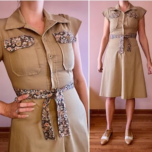 Può includere: Abito color beige con maniche corte, colletto e chiusura con bottoni sul davanti. L'abito presenta dettagli con stampa floreale sulle tasche sul petto e una cintura abbinata. L'abito è al ginocchio.