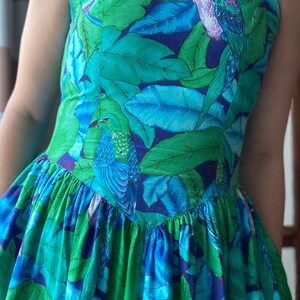Op de afbeelding: Een mouwloze jurk met een blauwe en groene tropische print met vogels en bladeren. De jurk heeft een getailleerd bovenlijf en een volle rok.
