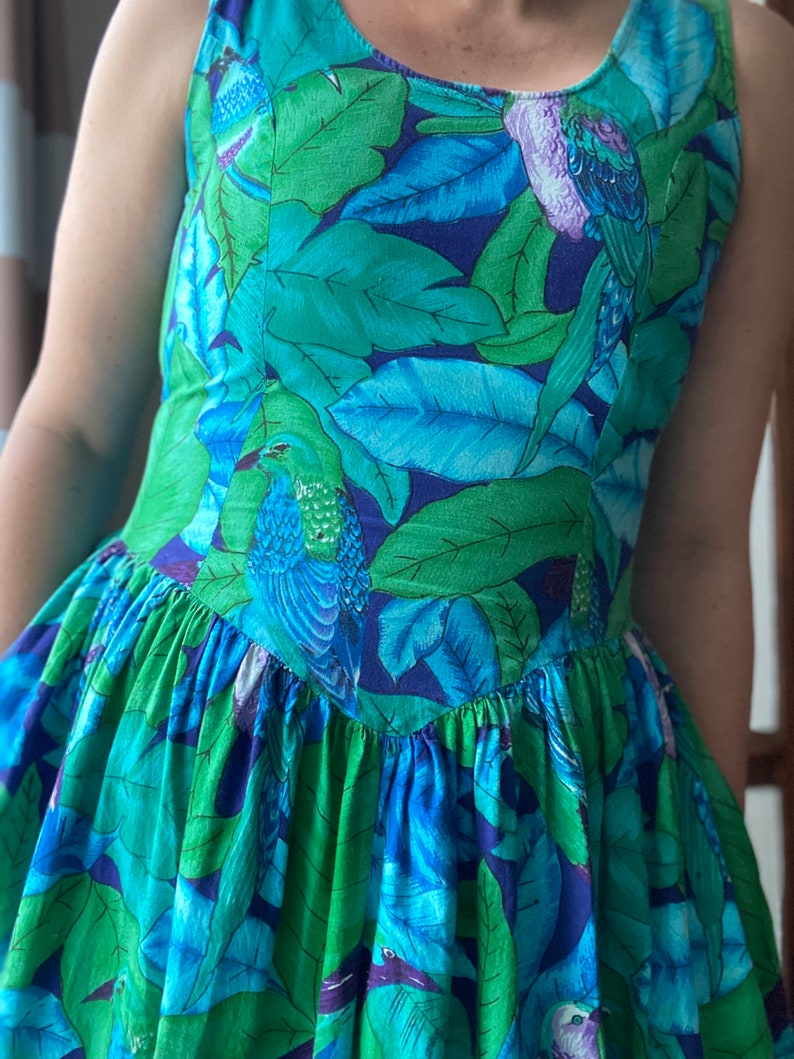 Op de afbeelding: Een mouwloze jurk met een blauwe en groene tropische print met vogels en bladeren. De jurk heeft een getailleerd bovenlijf en een volle rok.