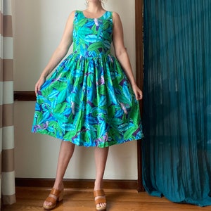 Op de afbeelding: Een vrouw draagt een blauwe en groene jurk met bloemenprint, een getailleerd bovenstuk en een volle rok. De jurk heeft een tropisch thema met bladeren en vogels.