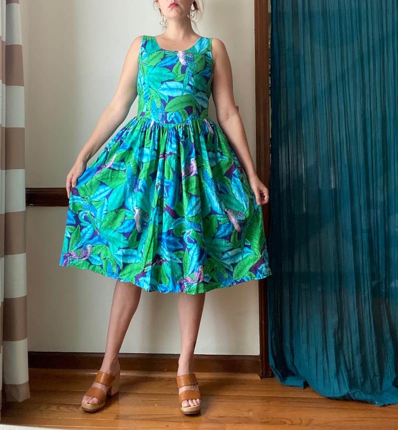 Op de afbeelding: Een vrouw draagt een blauwe en groene jurk met bloemenprint, een getailleerd bovenstuk en een volle rok. De jurk heeft een tropisch thema met bladeren en vogels.