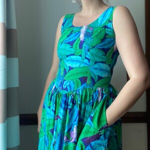 Op de afbeelding: Een mouwloze jurk met een blauwe en groene bloemenprint met vogels. De jurk heeft een getailleerd bovenstuk en een volle rok met zakken.