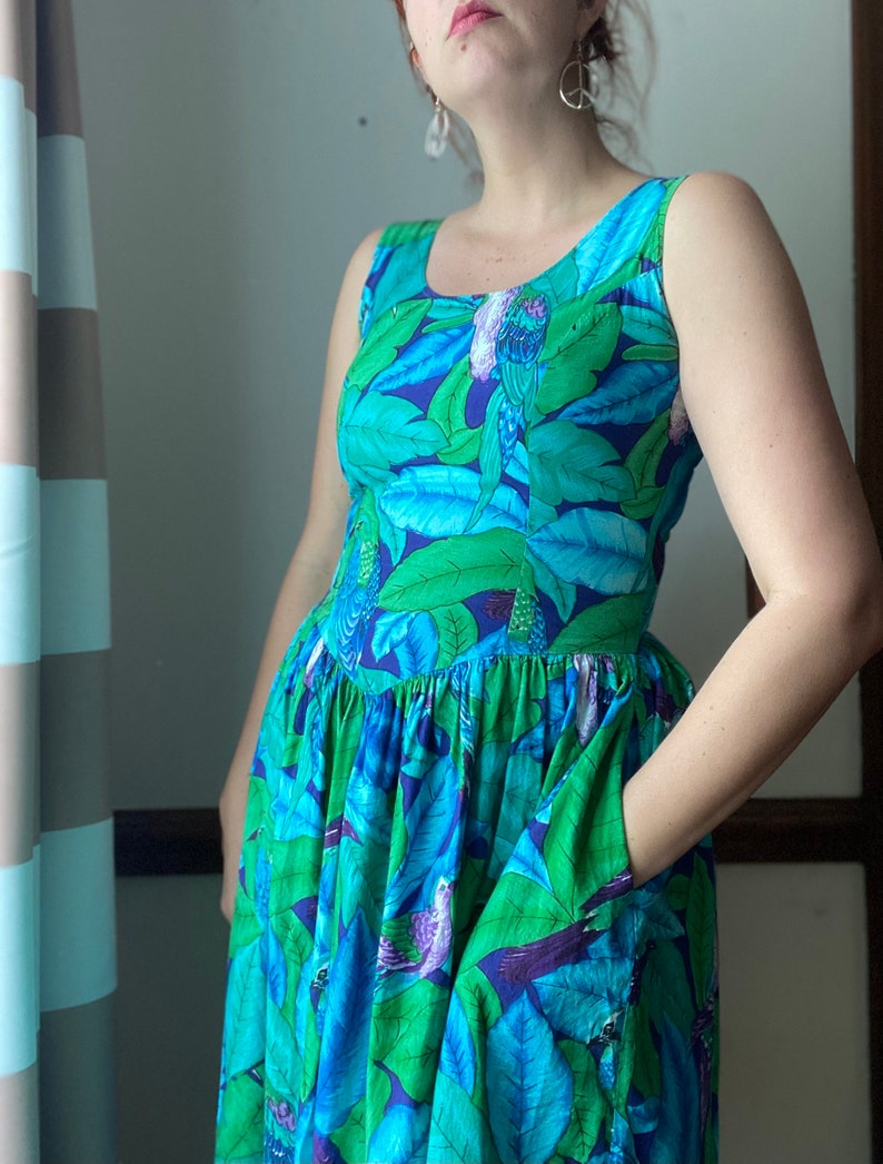 Op de afbeelding: Een mouwloze jurk met een blauwe en groene bloemenprint met vogels. De jurk heeft een getailleerd bovenstuk en een volle rok met zakken.