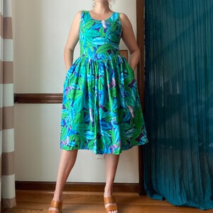 Op de afbeelding: Een vrouw draagt een blauwe en groene tropische printjurk met een mouwloos lijfje en een volle rok. De jurk heeft een bloemenpatroon met vogels.