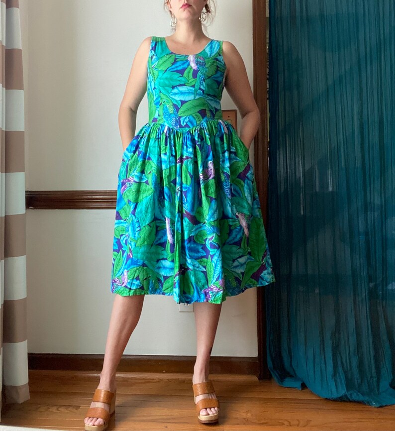 Op de afbeelding: Een vrouw draagt een blauwe en groene tropische printjurk met een mouwloos lijfje en een volle rok. De jurk heeft een bloemenpatroon met vogels.