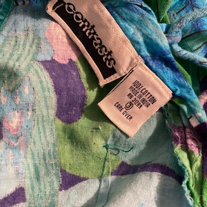 Op de afbeelding: Een wit label met zwarte tekst dat "Contrasts" zegt en een wit label met zwarte tekst dat "100% Katoen Gemaakt in India RN 31269 Verzorging op het label" zegt op een blauwe, groene en paarse bloemenstof.