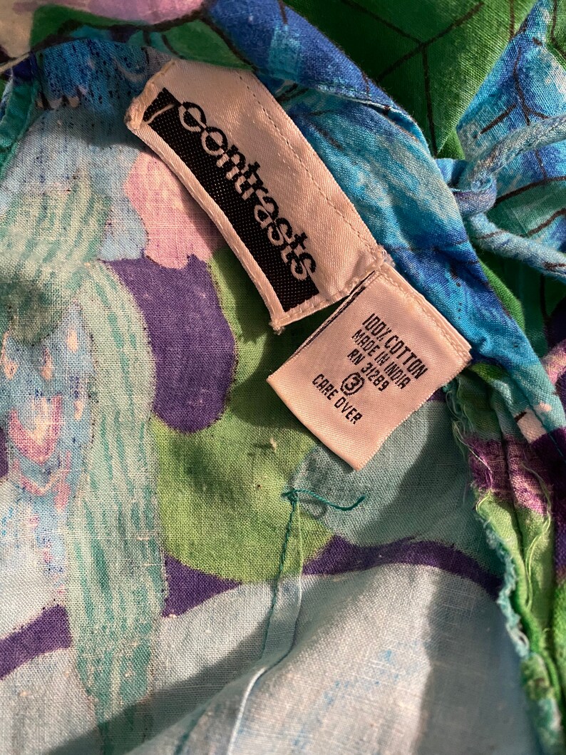 Op de afbeelding: Een wit label met zwarte tekst dat "Contrasts" zegt en een wit label met zwarte tekst dat "100% Katoen Gemaakt in India RN 31269 Verzorging op het label" zegt op een blauwe, groene en paarse bloemenstof.