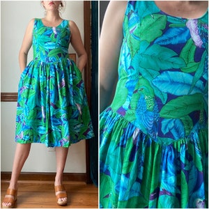 Op de afbeelding: Een blauwe en groene jurk met bloemenprint, een getailleerd bovenstuk en een volle rok. De jurk heeft een ronde halslijn en is mouwloos.